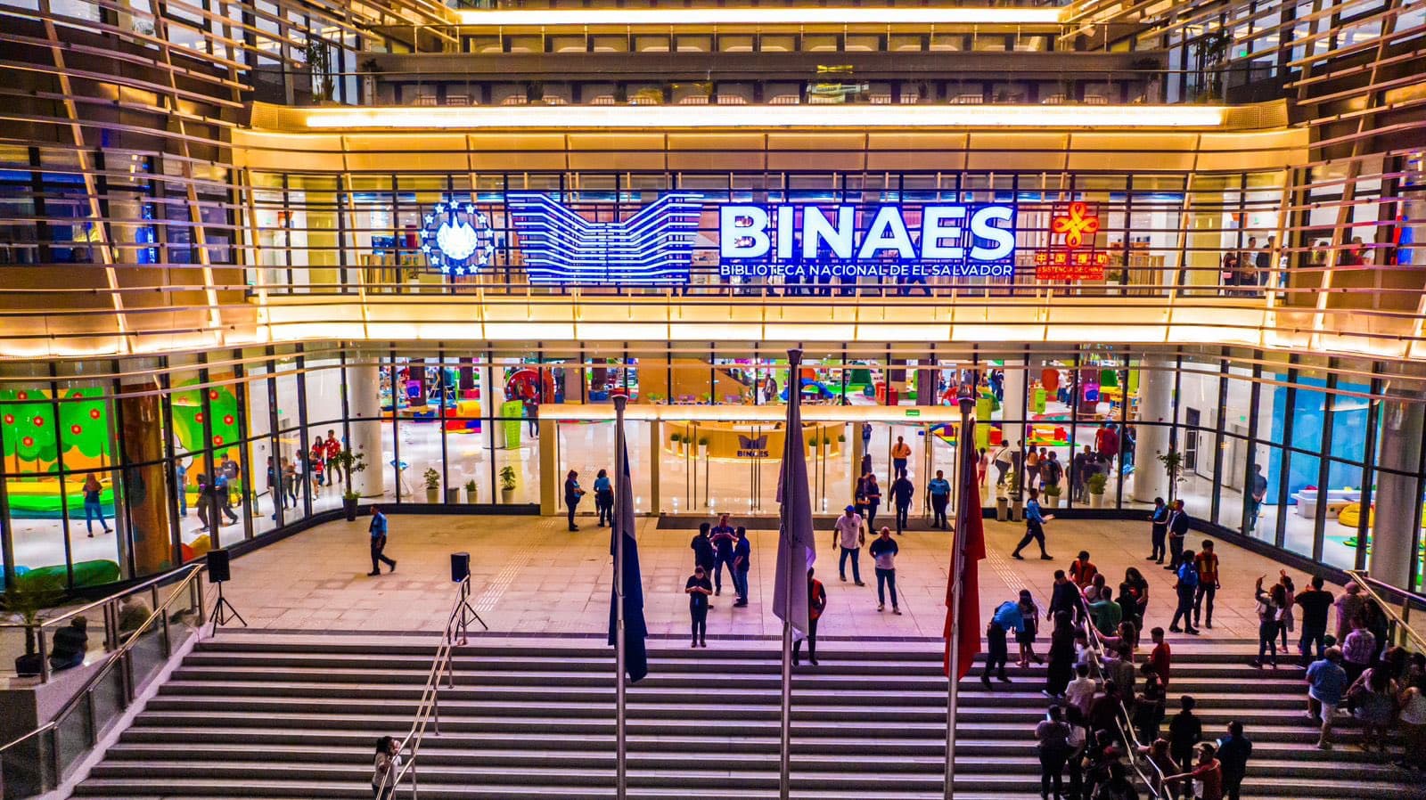 Modern glass facade of BINAES library in El Salvador illuminated at night with visitors.