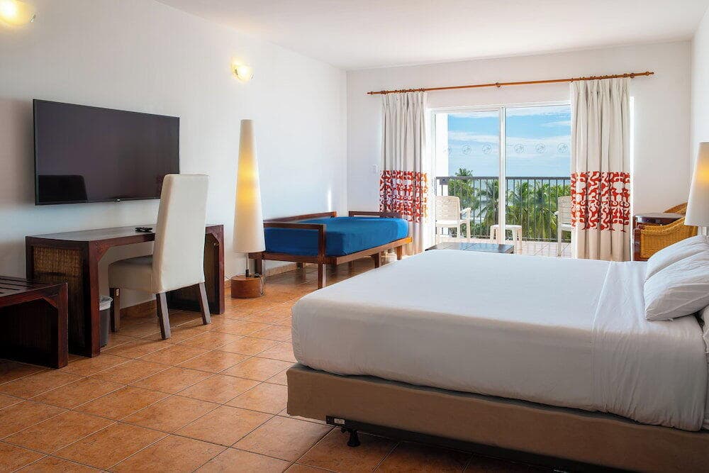 Bright hotel room with white bed, desk, blue daybed, and balcony overlooking tropical palm trees.