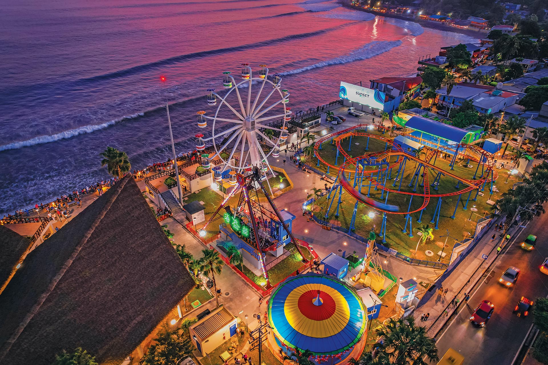 Aerial view of a brightly lit amusement park beside the ocean during a purple sunset.